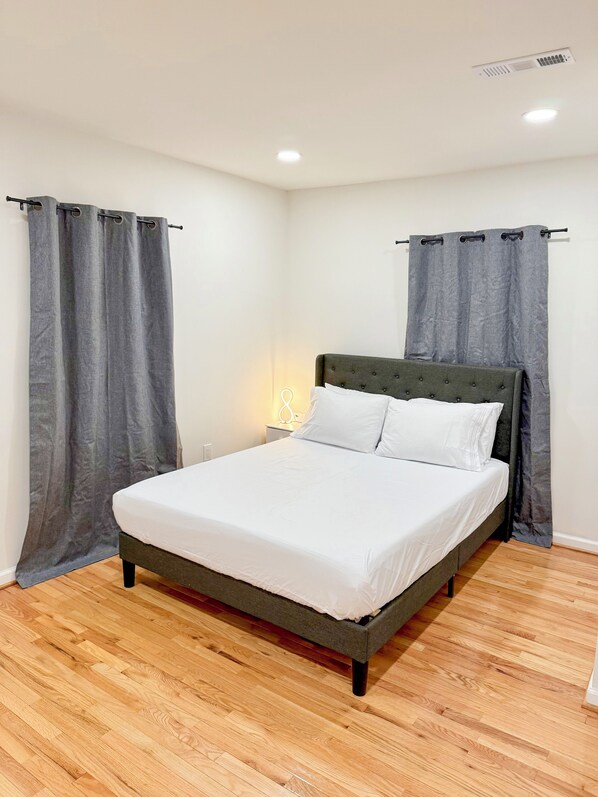 A bedroom with a bed, grey curtains, a bedside lamp, and a wooden floor.
