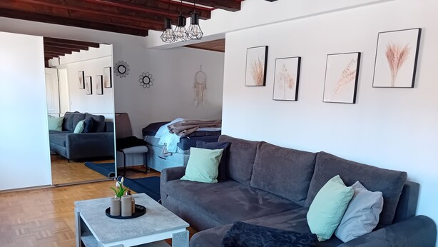 Ein modernes Wohnzimmer mit einem grauen Sofa, einem Couchtisch und einem Spiegel. An den Wänden befinden sich gerahmte Bilder und ein Traumfänger.