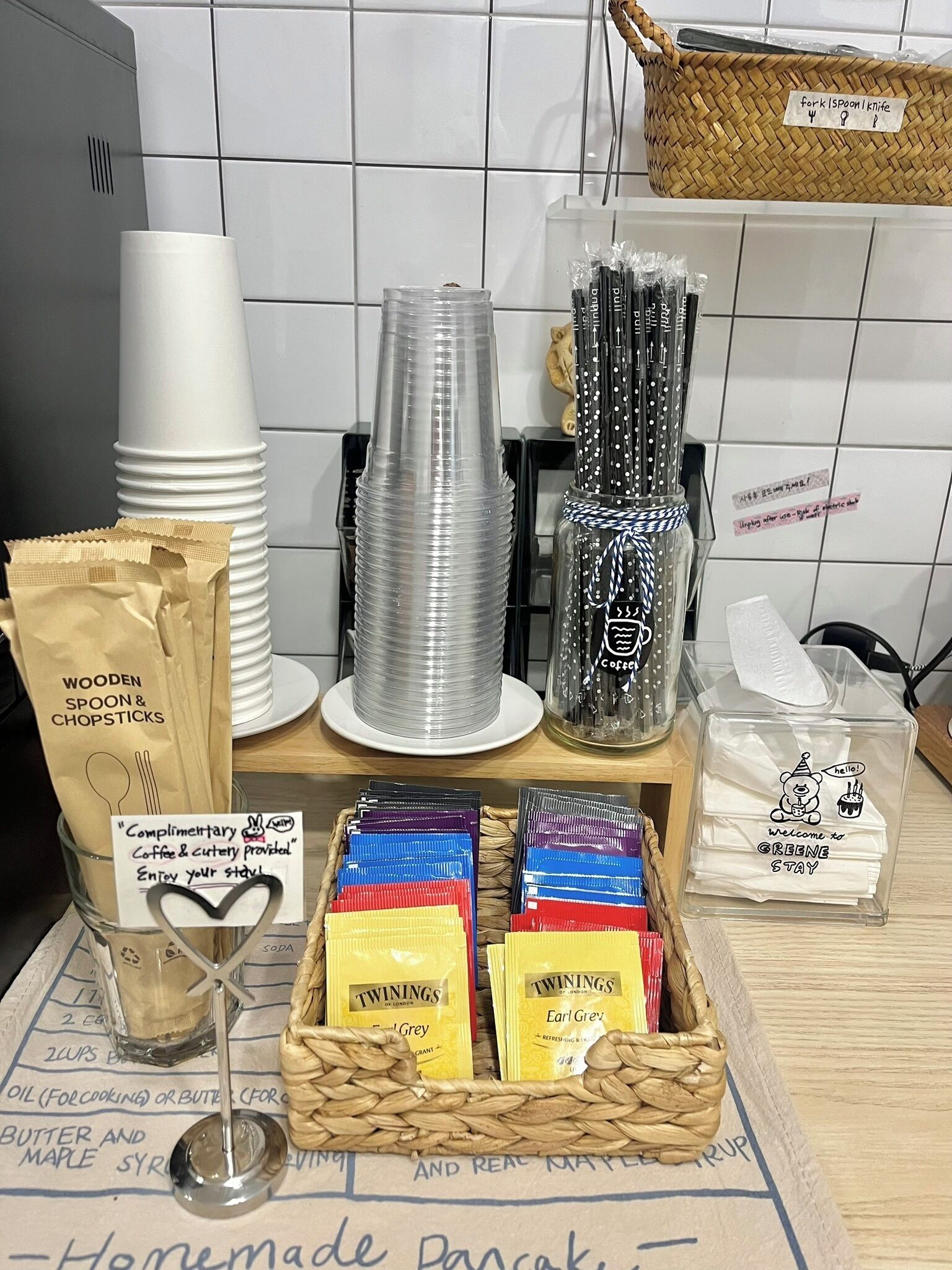 A kitchen counter with various utensils and food items, including stacked plastic cups, a basket of wooden spoon and chopsticks, a jar of coffee, and boxes of Twinings Earl Grey tea.