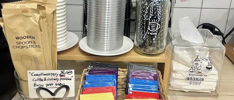 A kitchen counter with various utensils and food items, including stacked plastic cups, a basket of wooden spoon and chopsticks, a jar of coffee, and boxes of Twinings Earl Grey tea.