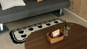 A modern living room with a grey sofa, a mustard-colored pillow, a white textured pillow, a dark brown coffee table with a basket and a candle, and a black and white patterned rug. - GreeneStay (Seoul)