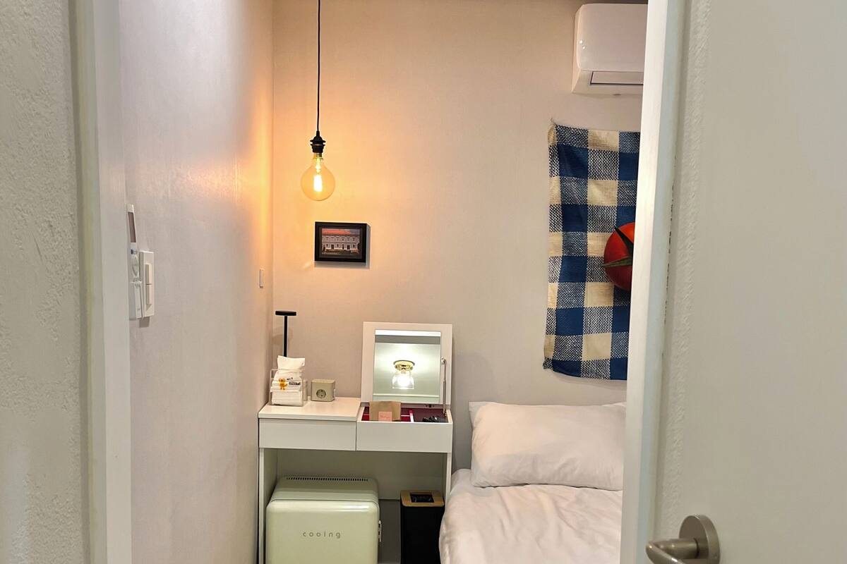 A narrow hotel room with a bed, a desk with a computer, a small table with a basket, and a wall-mounted air conditioner.