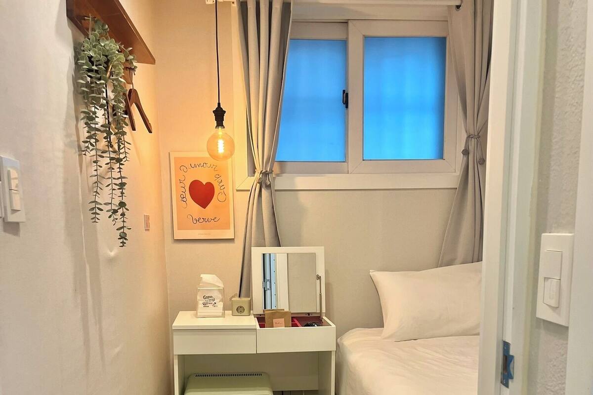 A compact bedroom with a bed, a bedside table with a lamp, a small desk with a computer, a radiator, and a window with curtains.