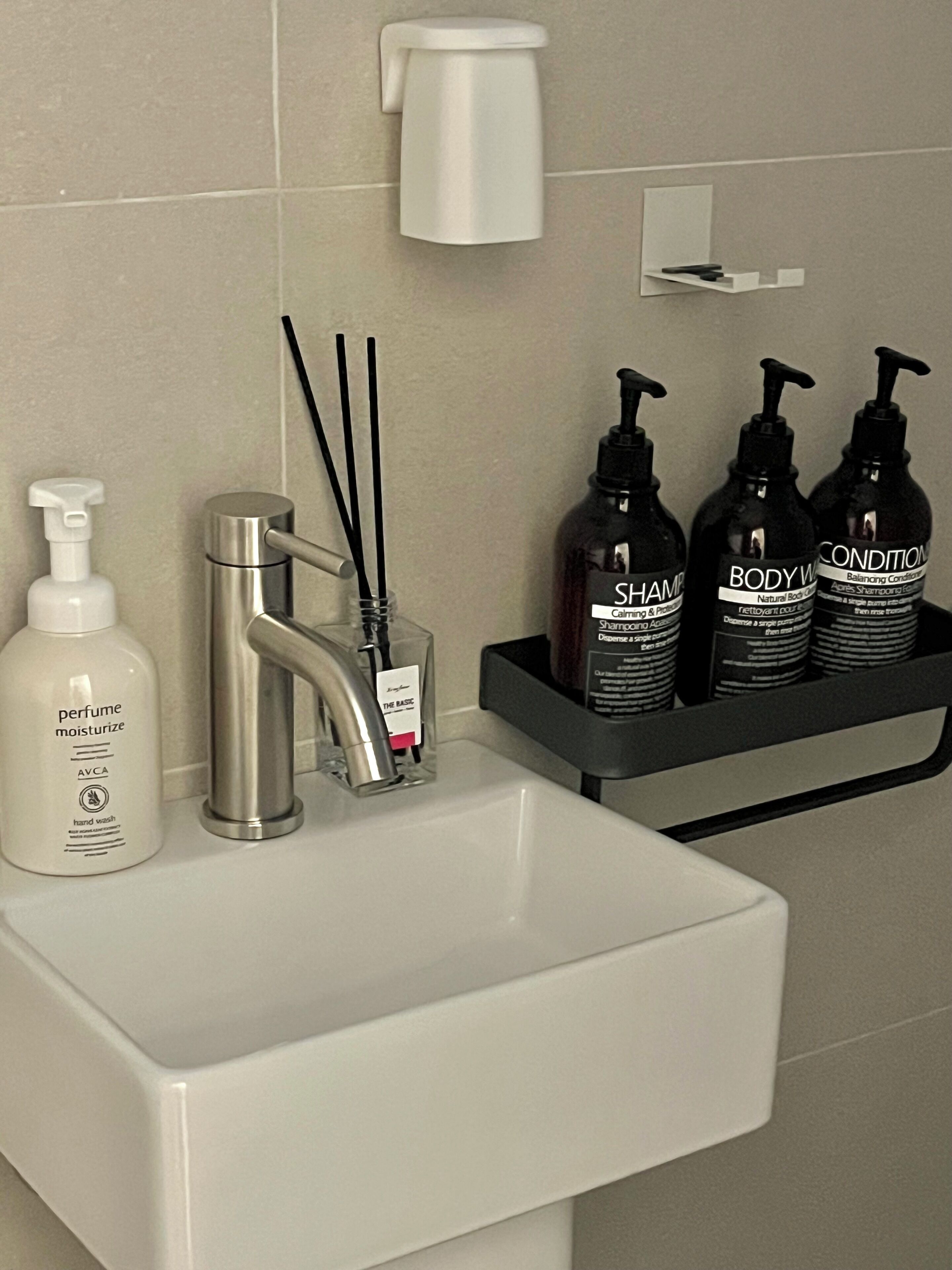 Un baño con un lavamanos blanco, un grifo plateado, un dispensador de jabón y tres botellas negras de gel de ducha sobre un estante negro.