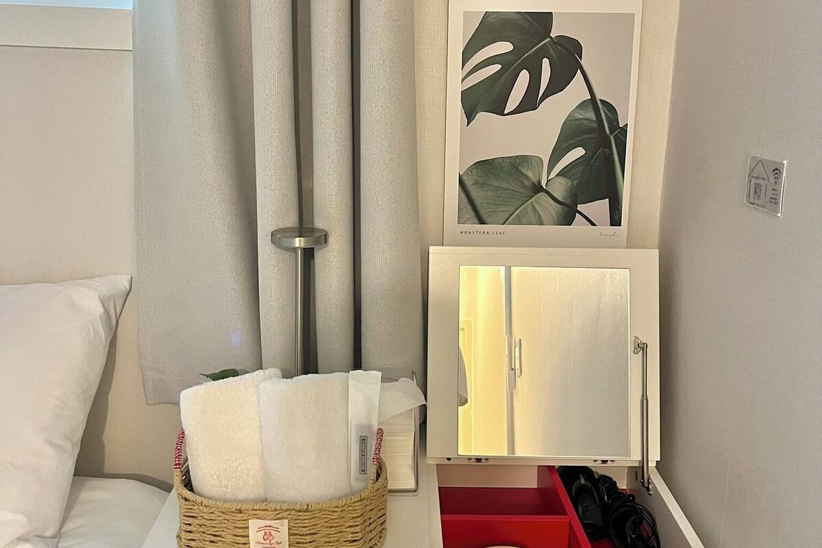 A neatly organized bedroom with a bed, a bedside table with a basket, a framed plant, a mirror, and a hanging light fixture.
