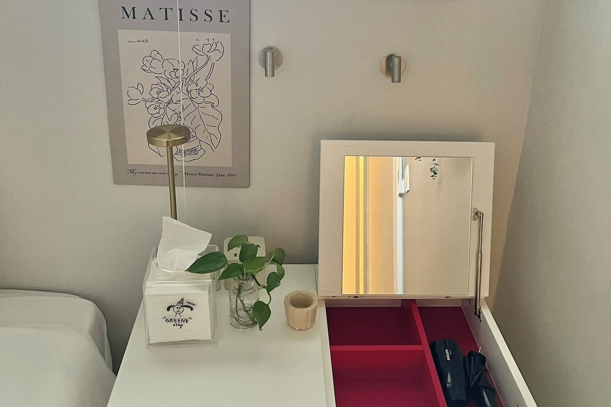 A white bedside table with a red drawer, a framed artwork of Matisse, a small plant, and a light bulb hanging above.