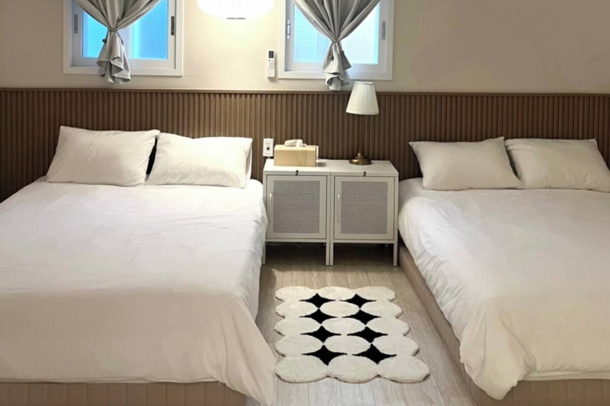 A hotel room with two beds, a small bedside table with a lamp, a decorative rug, and a window with curtains.