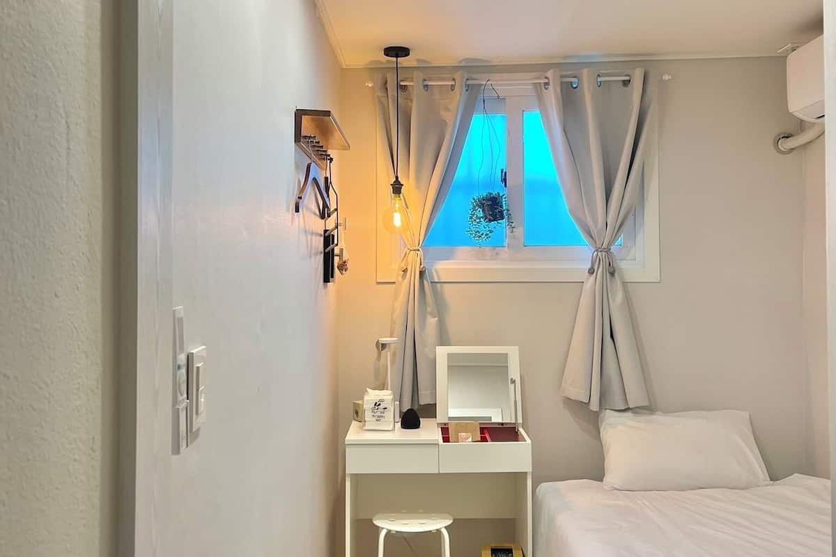 A bedroom with a bed, a desk with a chair, a small stool, a mirror, a lamp, and a window with curtains.