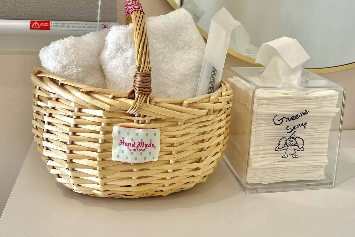 A wicker basket with a tag labeled "Hand Made with Love" and a clear plastic container with white napkins labeled "Greene Stay."