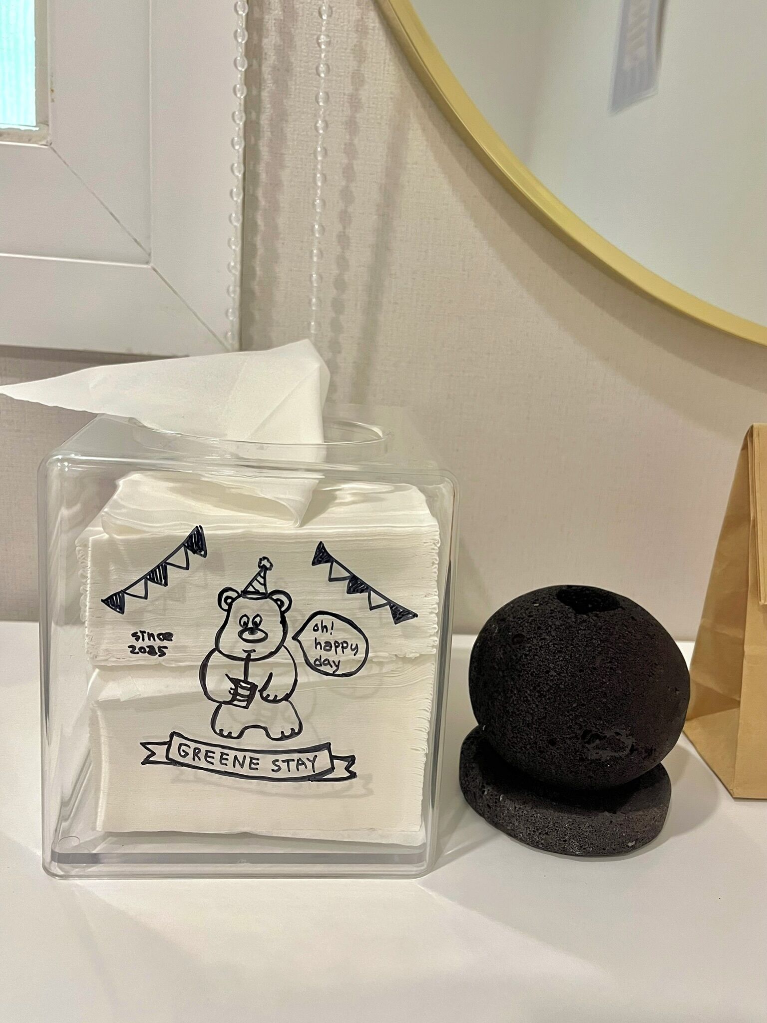 A clear glass candle with a bear illustration and text, a black stone, and a brown paper bag.