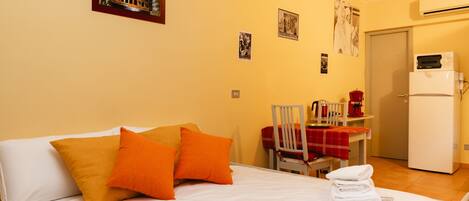 A hotel room with a bed, pillows, a chair, a table, a refrigerator, and a wall with pictures.