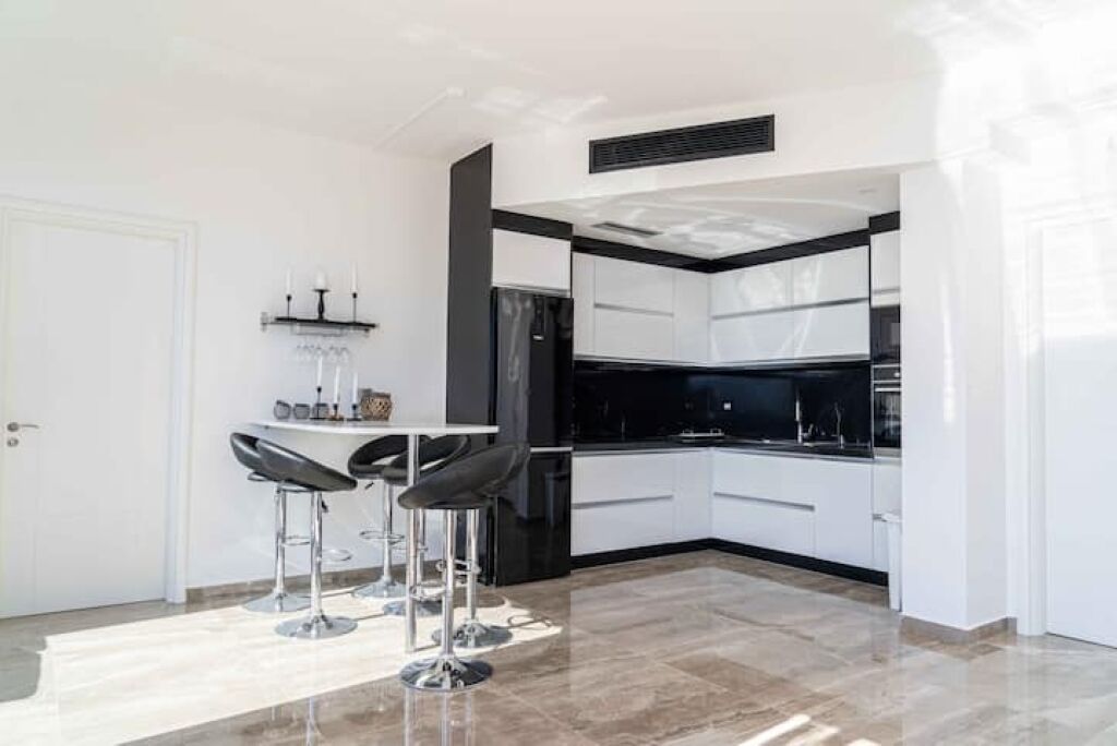 Una cucina moderna con mobili bianchi, un piano di lavoro nero e un forno integrato. Ci sono due sgabelli da bar e un piccolo tavolo con un vaso.