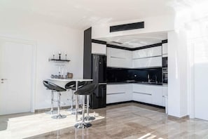 A modern kitchen with white cabinets, a black countertop, and a built-in oven. There are two bar stools and a small table with a vase.