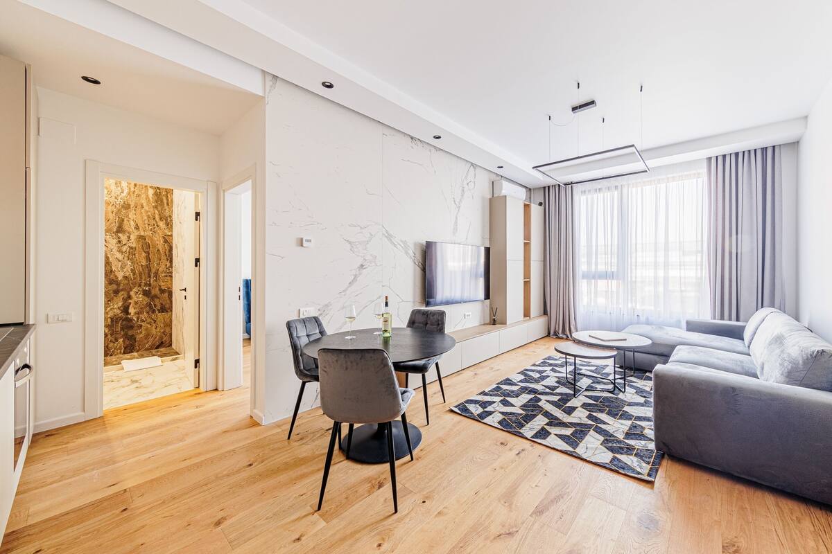 A modern living room with a dining area, a grey sofa, a patterned rug, and a TV mounted on the wall.