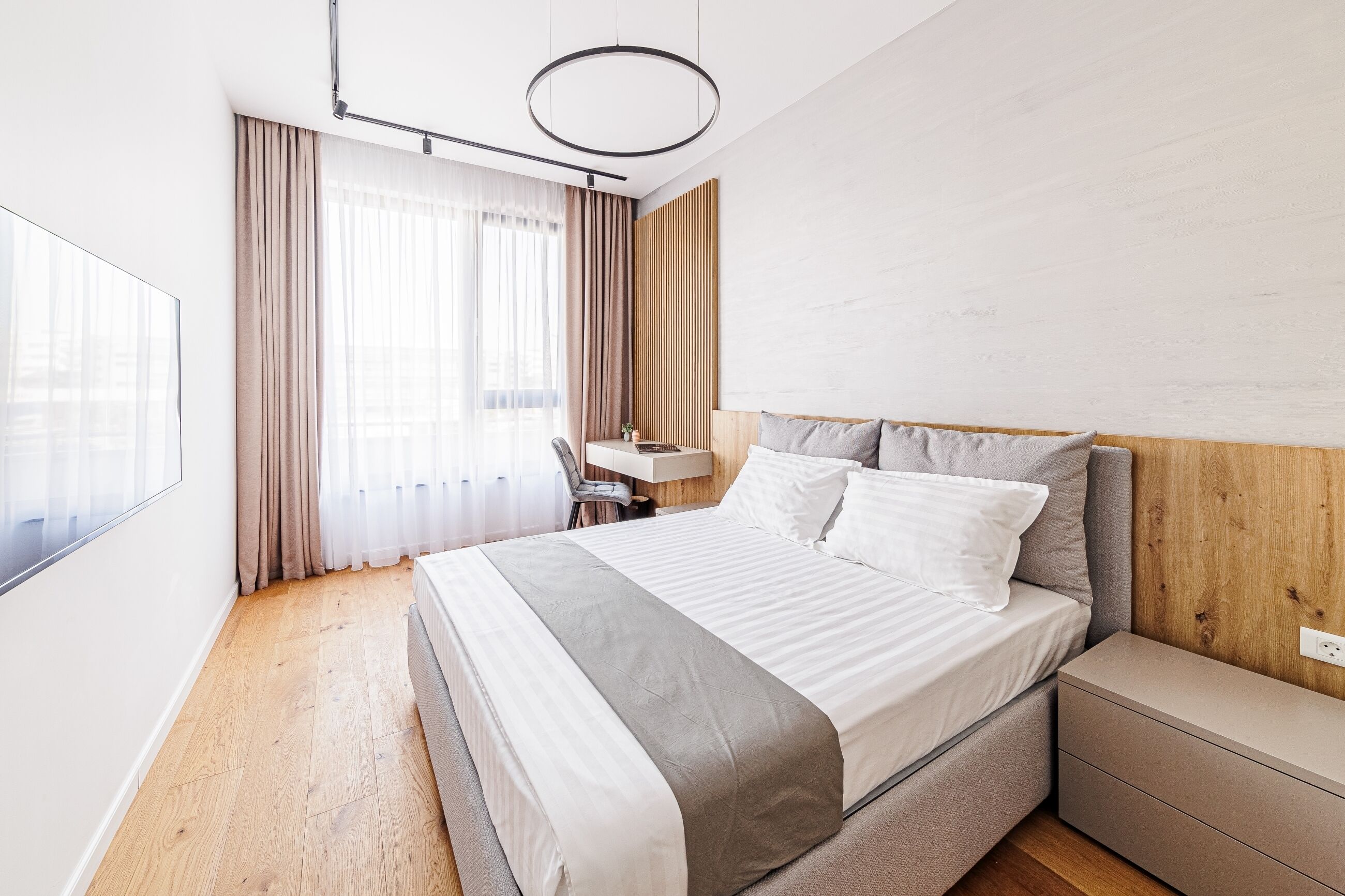 Una habitación de hotel moderna con una cama grande, una mesita de noche, un escritorio y un amplio ventanal con cortinas.