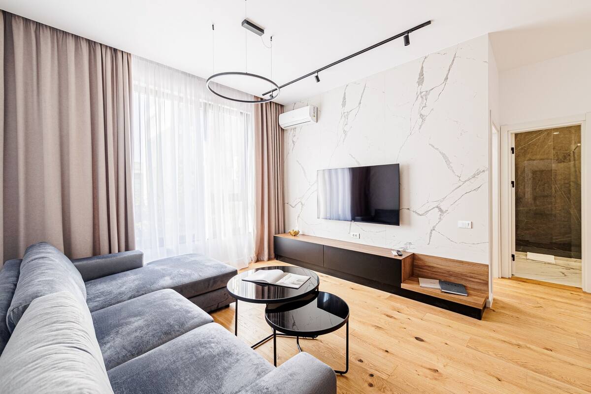 A modern living room with a sectional sofa, a round black coffee table, and a flat-screen TV mounted on a marble wall.