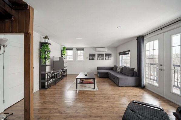 A spacious living room with a wooden floor, a grey sofa, a coffee table, and a TV on a stand. There are large windows on one side and a door on the other. - Escape to Grandes-Piles (Grandes-Piles)