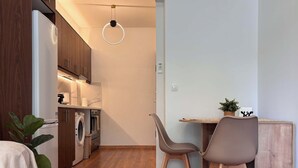 Kompakti keittiö, jossa on modernit kodinkoneet, ruokailutila kahdella tuolilla ja puulattia.