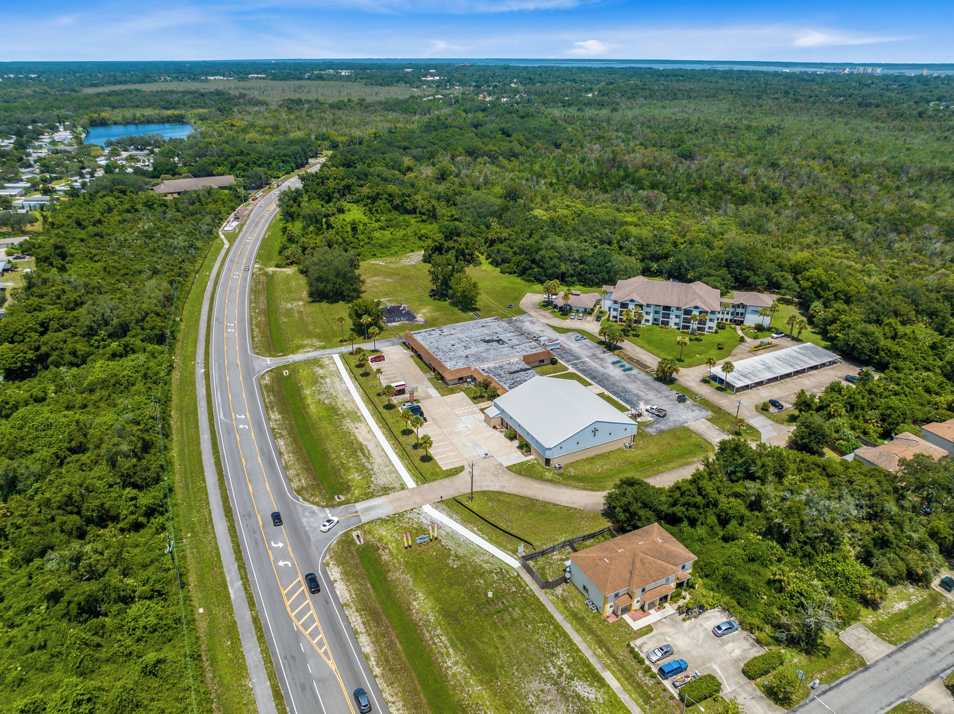 Une vue aérienne d’une zone commerciale avec un grand parking, un bâtiment doté d’un toit plat et une route qui serpente à travers le paysage.