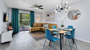 Un salon moderne avec un canapé d’angle marron, une table à manger avec des chaises bleues, un ventilateur de plafond et une grande fenêtre ornée de rideaux turquoise.