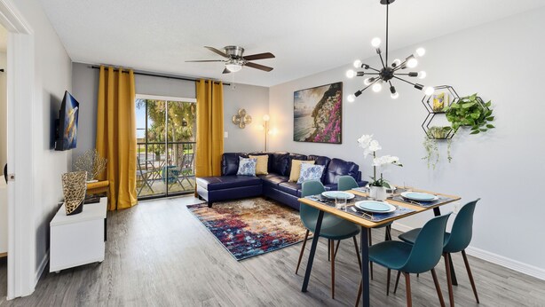 Un salon moderne avec un canapé d’angle, une table à manger avec des chaises, un ventilateur de plafond et une grande fenêtre ornée de rideaux jaunes.