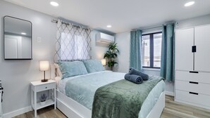 Une chambre à coucher comprenant un lit, une table de chevet, un miroir, un appareil de climatisation et une fenêtre avec des rideaux.