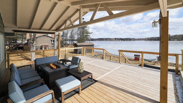 Une terrasse couverte avec des sièges d’extérieur et un foyer, offrant une vue sur un lac et une plage.