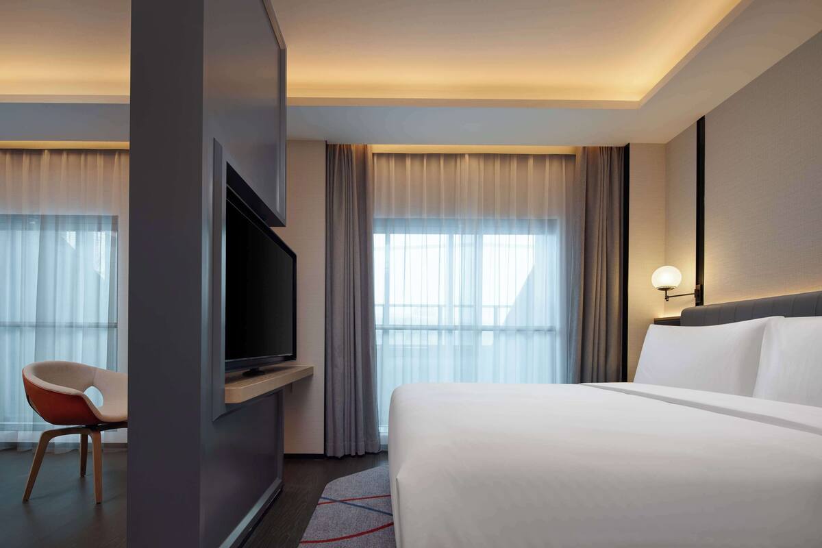 Una habitación de hotel con cama, televisión sobre un mueble, una silla y una ventana con cortinas.