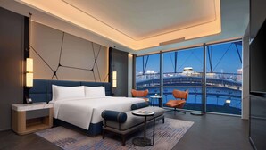 Una habitación de hotel moderna con una cama grande, un área de descanso con una mesa y vistas a la ciudad a través de grandes ventanales.