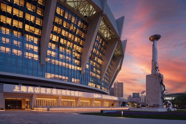 A modern building with a curved facade and large windows, featuring a prominent sculpture in the foreground. - Radisson Blu Hotel, Shanghai Stadium (Shanghai)