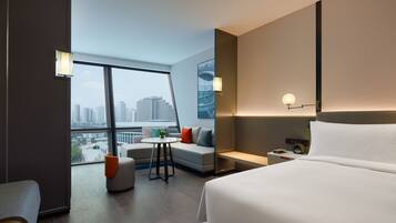 Habitación de hotel moderna con una cama grande, un área de descanso con una mesita y vistas a la ciudad a través de amplias ventanas.