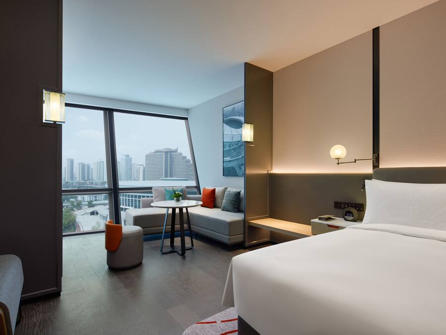 Habitación de hotel moderna con una cama grande, un área de descanso con una mesita y vistas a la ciudad a través de amplias ventanas.