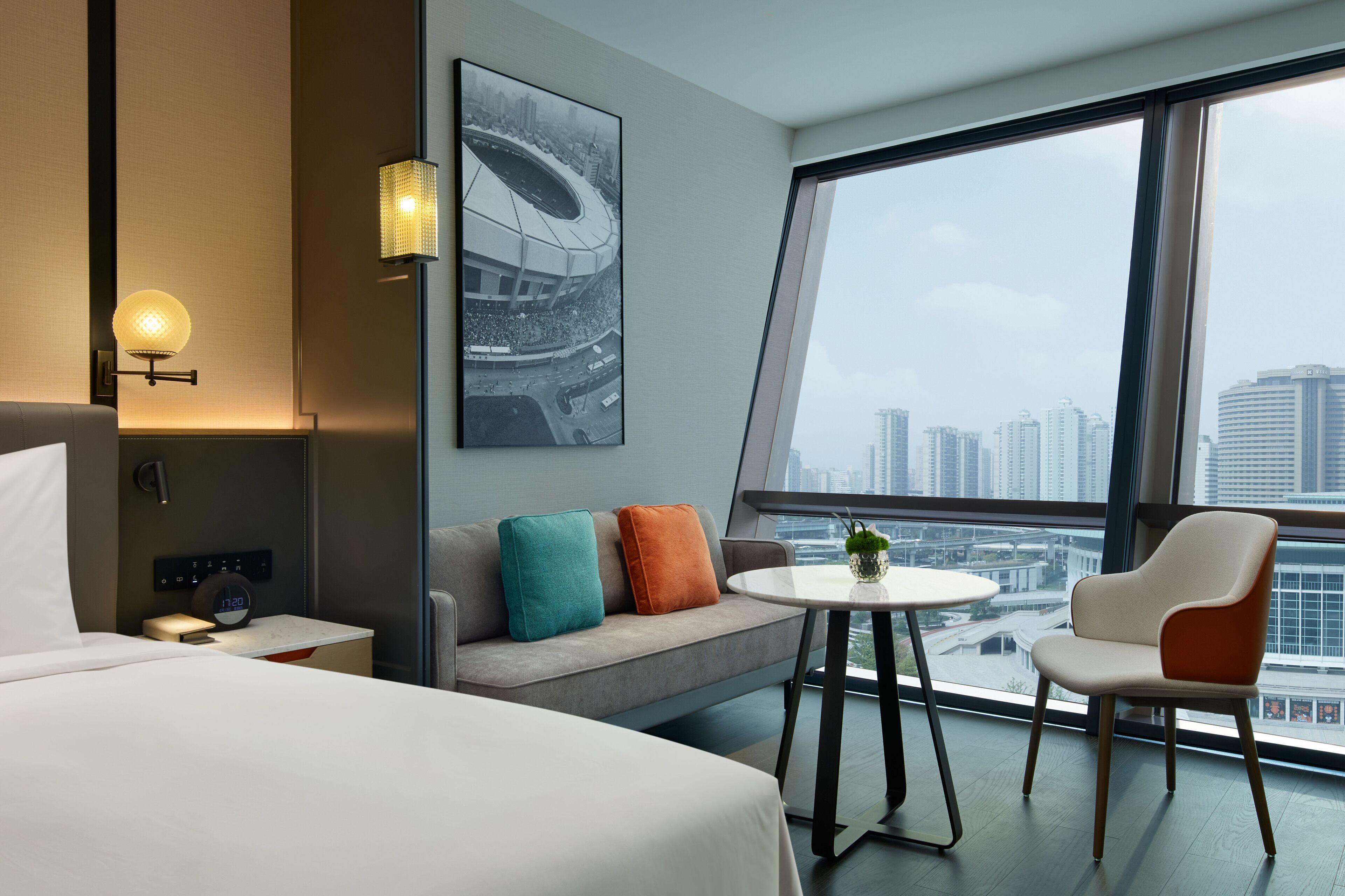 A modern hotel room with a bed, a sofa with colorful cushions, a small table with a plant, and a view of a cityscape through large windows.