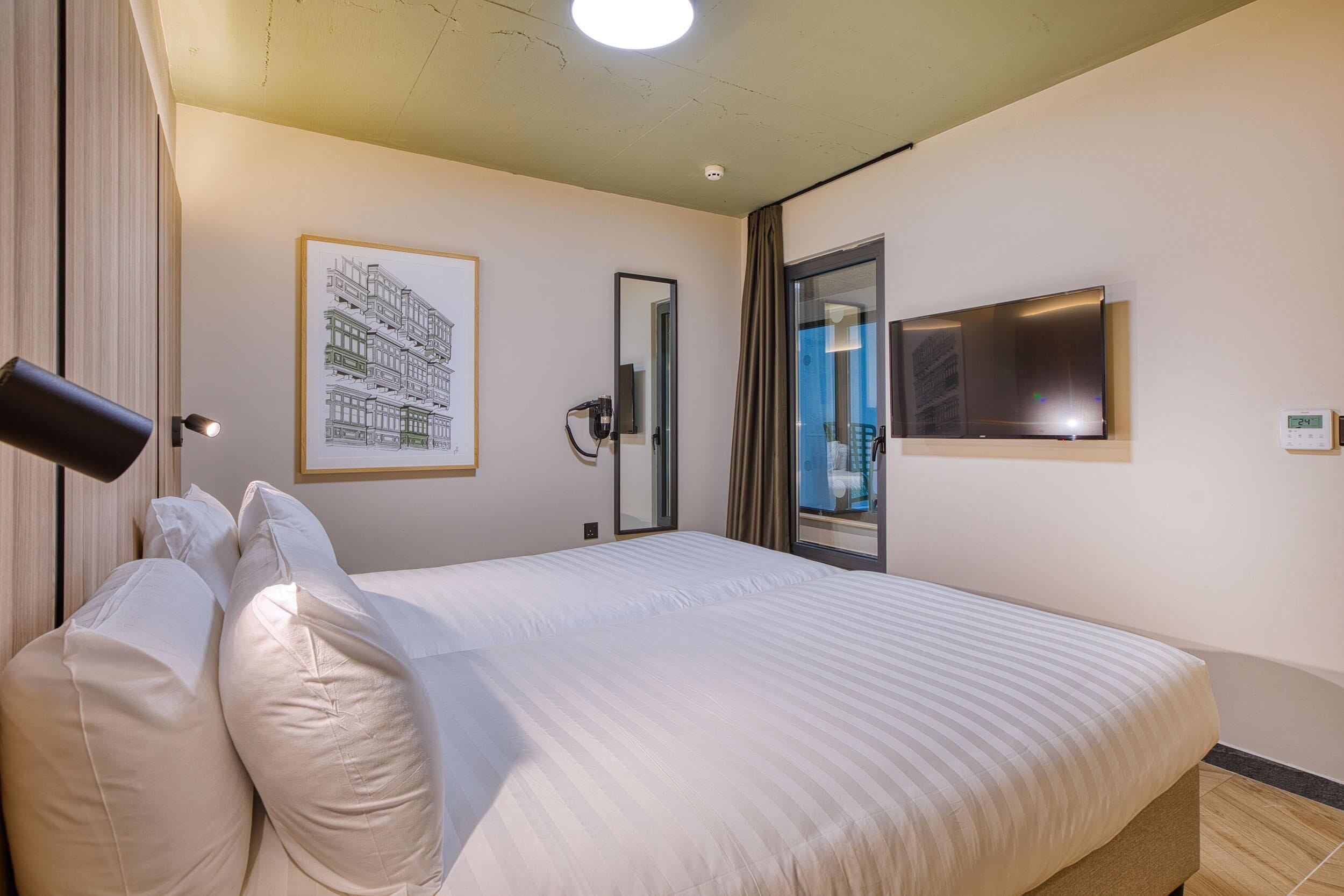 Ein Hotelzimmer mit einem großen Bett, einem an der Wand befestigten Fernseher, einem gerahmten Bild und einer Tür zu einem Balkon.