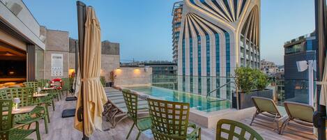 A rooftop bar with green chairs and a pool, surrounded by modern buildings.