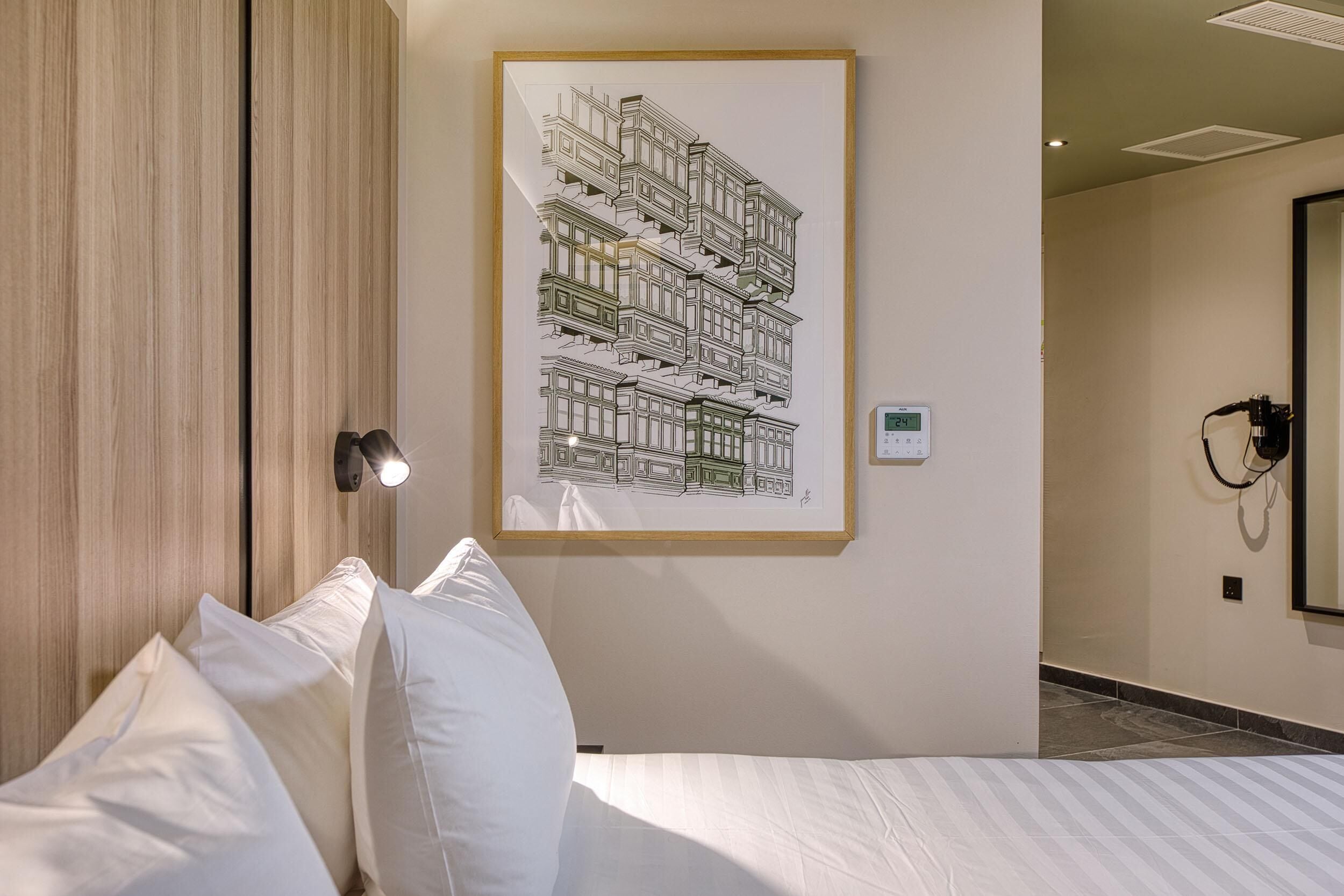 A hotel room with a bed, white pillows, a framed architectural drawing on the wall, a bedside lamp, and a thermostat.
