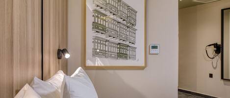 A hotel room with a bed, white pillows, a framed architectural drawing on the wall, a bedside lamp, and a thermostat.