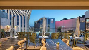 An outdoor dining area with modern chairs and tables, a view of the cityscape, and a building with a unique facade. - VITA Hotel (St. Julian's)