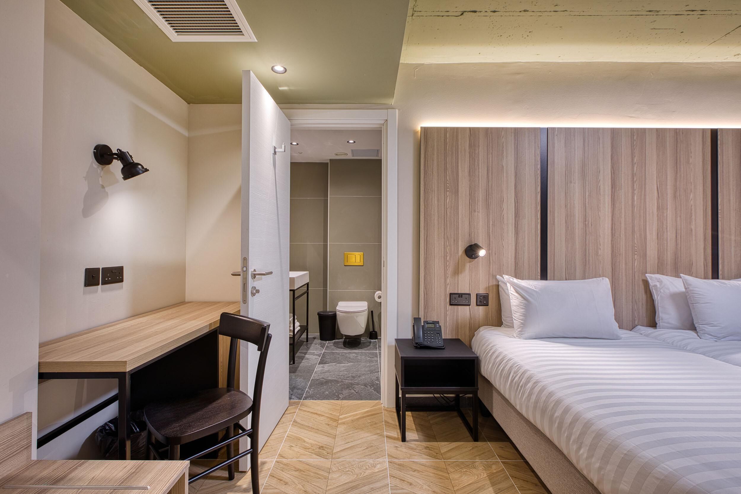 Ein modernes Hotelzimmer mit einem Holztisch, einem Bett mit weißer Bettwäsche, einem Nachttisch mit Telefon und einem Holzvorhang.