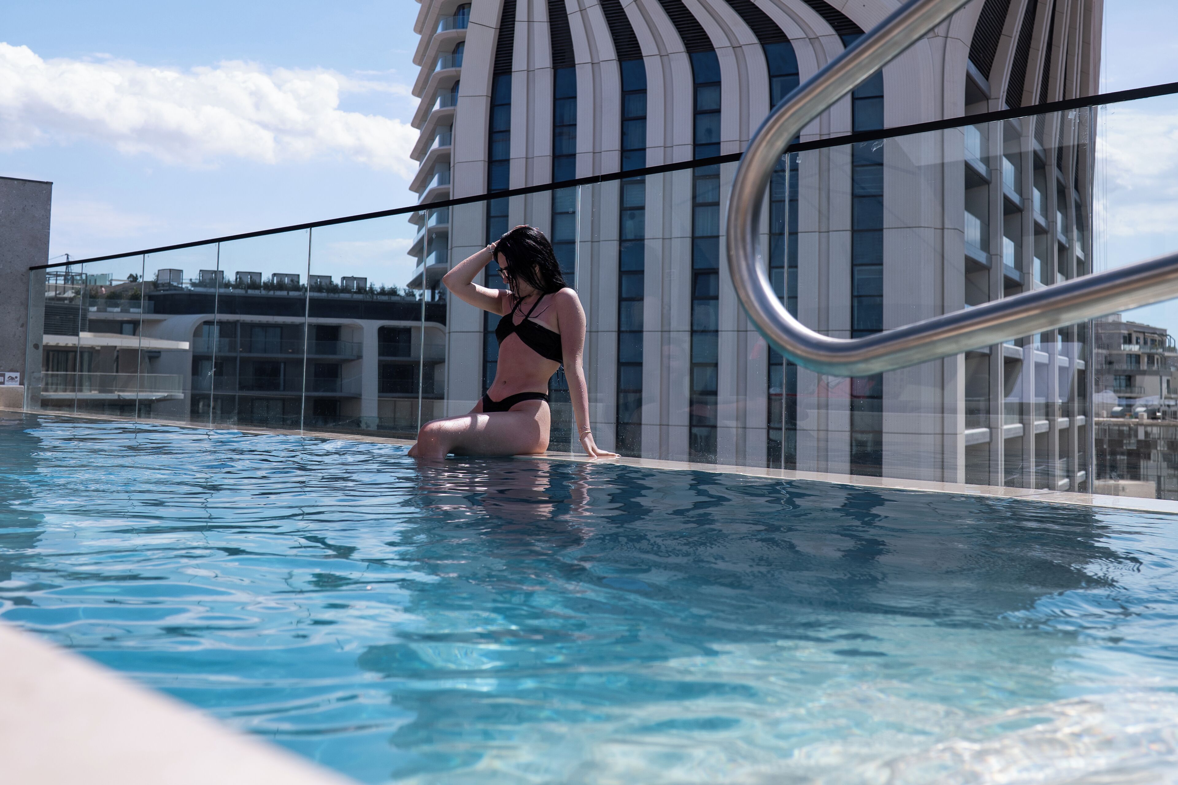 Une femme vêtue d’un bikini noir est assise au bord d’une piscine, avec un bâtiment moderne en arrière-plan.