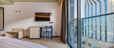 Una habitación de hotel moderna con una cama grande, un escritorio y una silla. Hay un televisor montado en la pared y una ventana con vistas a un edificio.