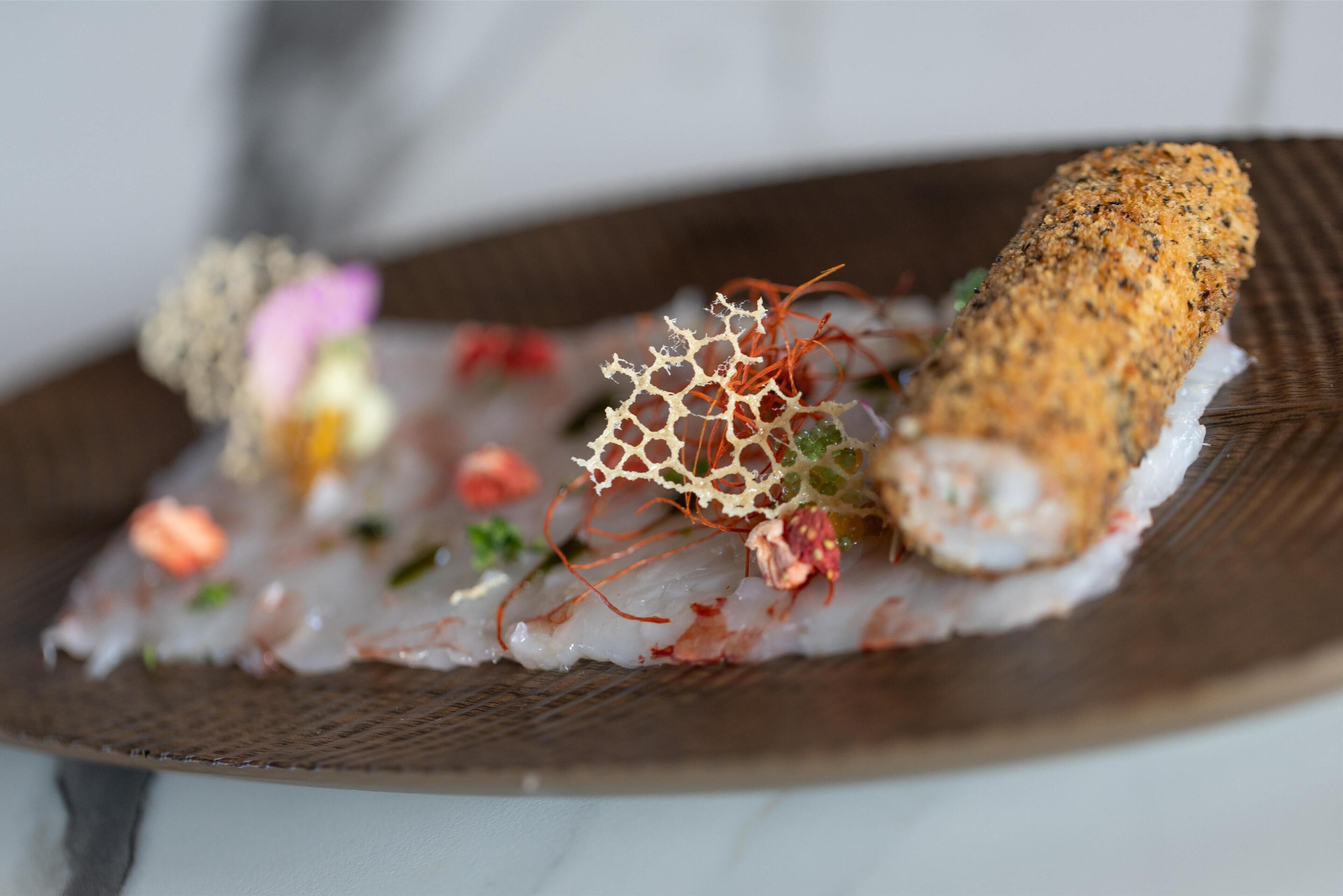 Un plato de sushi con una capa crujiente, decorado con alga marina delicada y verduras coloridas sobre un plato de madera oscura.