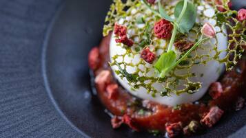 Un plat composé d’un élément blanc en forme de sphère, agrémenté de feuilles vertes et d’éléments rouges, le tout présenté sur une assiette sombre.