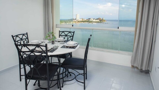 Un comedor con una mesa dispuesta para cuatro, una planta en maceta y vistas a una ciudad costera a través de amplios ventanales.