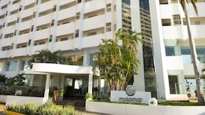 Un hotel de gran altura con varios pisos, un eje vertical de ascensor de vidrio y un área de entrada ajardinada con palmeras.