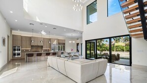 A modern kitchen with a central island, a dining area with chairs, and a large window offering a view of the outdoors. - SOL HAUS! LUXE NEW 6 BDRM BEACH HOUSE W/ STUNNING VIEWS/POOL/SPA/PUTTING GRN/GYM (Juno Beach)