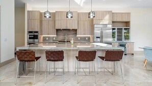 A modern kitchen with a central island, bar stools, stainless steel appliances, and pendant lighting. - SOL HAUS! LUXE NEW 6 BDRM BEACH HOUSE W/ STUNNING VIEWS/POOL/SPA/PUTTING GRN/GYM (Juno Beach)