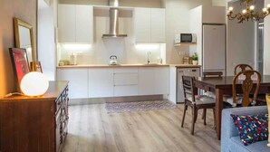 Une cuisine moderne avec des armoires blanches, une hotte de cuisinière en acier inoxydable et un four à micro-ondes. On y trouve un coin repas avec une table et des chaises en bois, ainsi qu’un tapis coloré sur le sol.