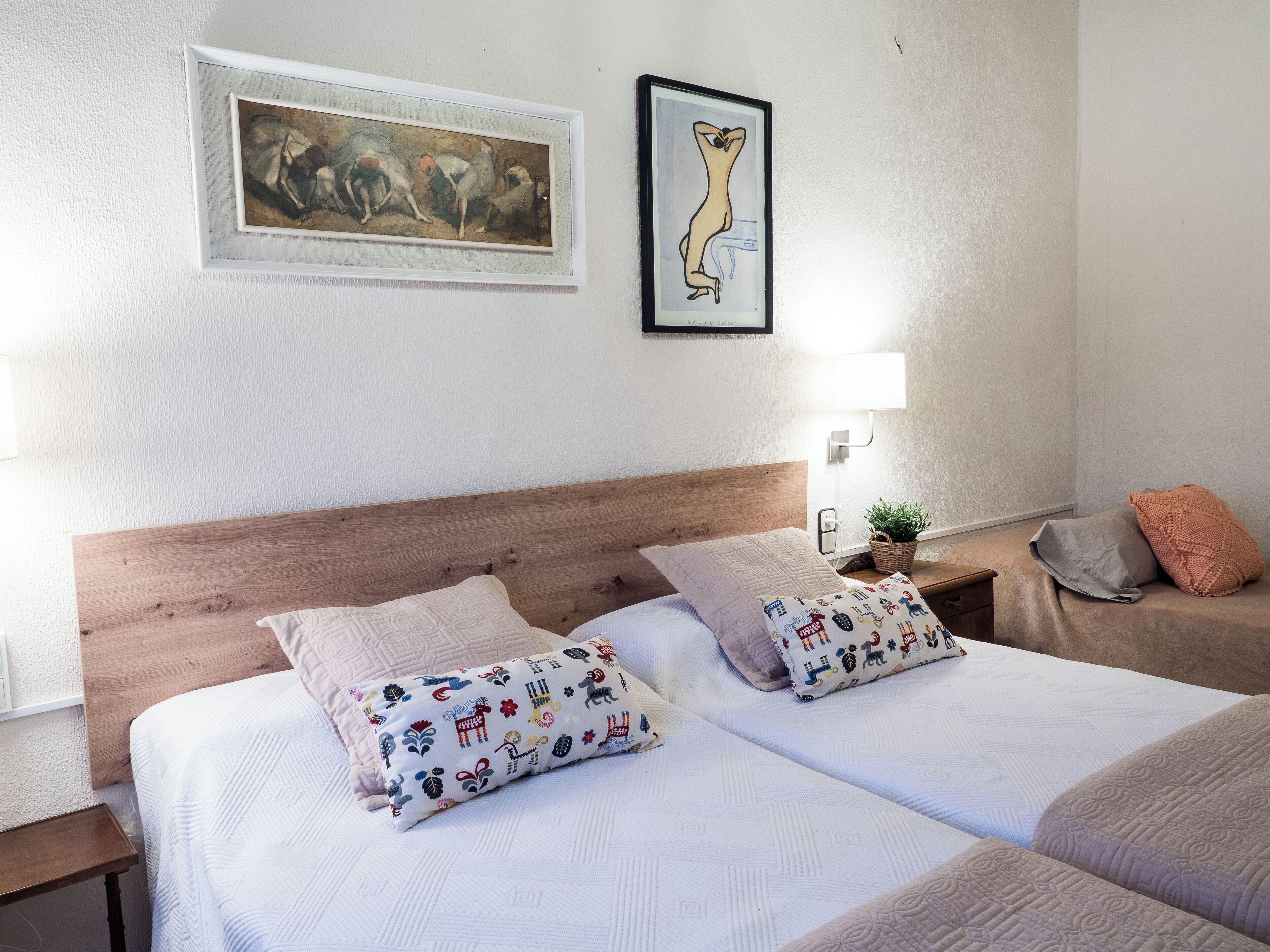 Een tweepersoonsbed met gestreepte kussens, een houten hoofdbord, een nachtkastje met een lamp en een plant, en twee ingelijste kunstwerken aan de muur.