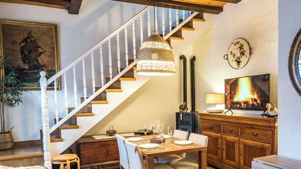 Een eetgedeelte met een houten tafel gedekt voor vier personen, een trap met witte spijlen, een hanglamp en een televisie waarop een scène van een haard te zien is.